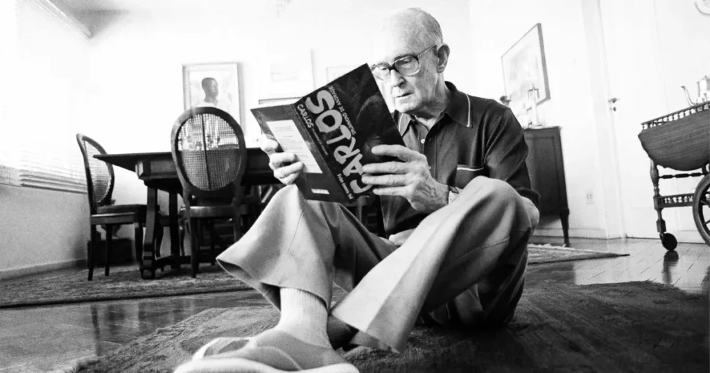 Carlos Drummond de Andrade sentado no chão de casa lendo um livro, em uma foto em preto e branco. Ícone da poesia brasileira e autor de obras como A Rosa do Povo e Claro Enigma.