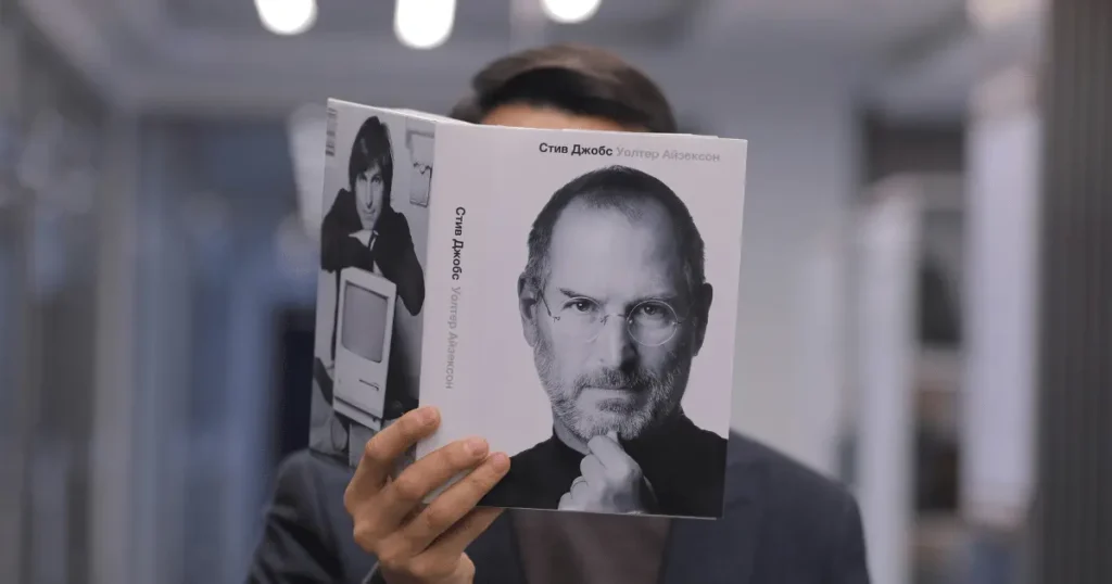 Pessoa segura o livro de Steve Jobs escrito por Walter Isaacson, com a capa alinhada ao rosto como se fosse o próprio Jobs — uma biografia famosa entre os melhores livros de biografias.