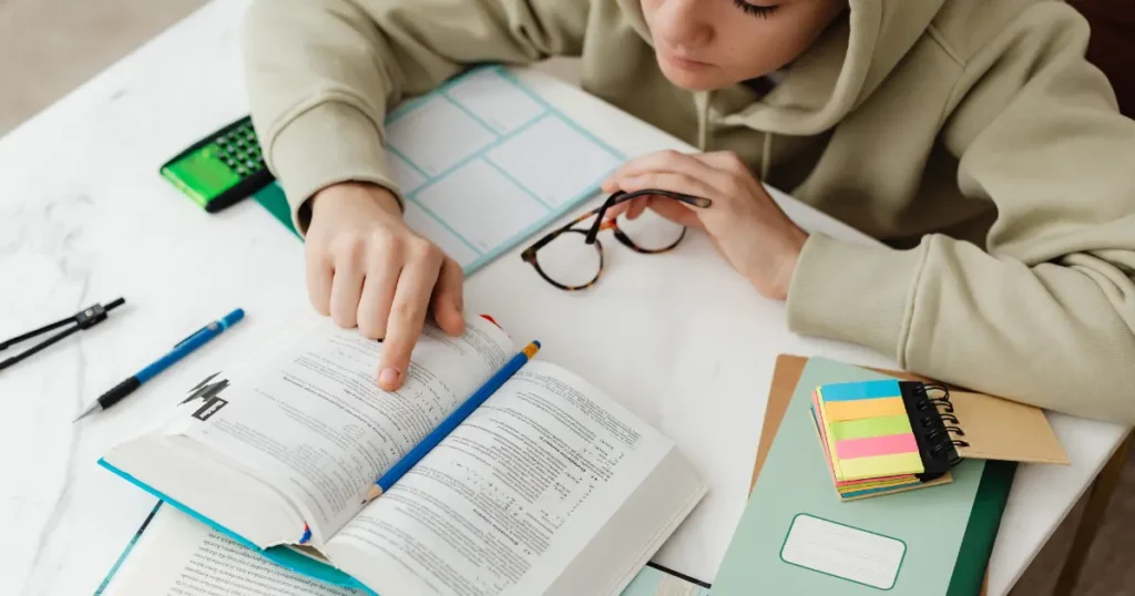 Estudante consultando um livro aberto em meio a materiais de estudo, ilustrando de forma prática o uso cotidiano de livros didáticos.